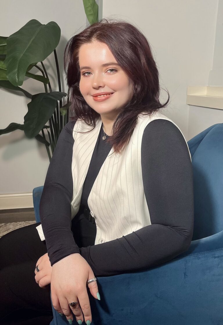 Maeve Kell smiling in a blue armchair beside a large plant