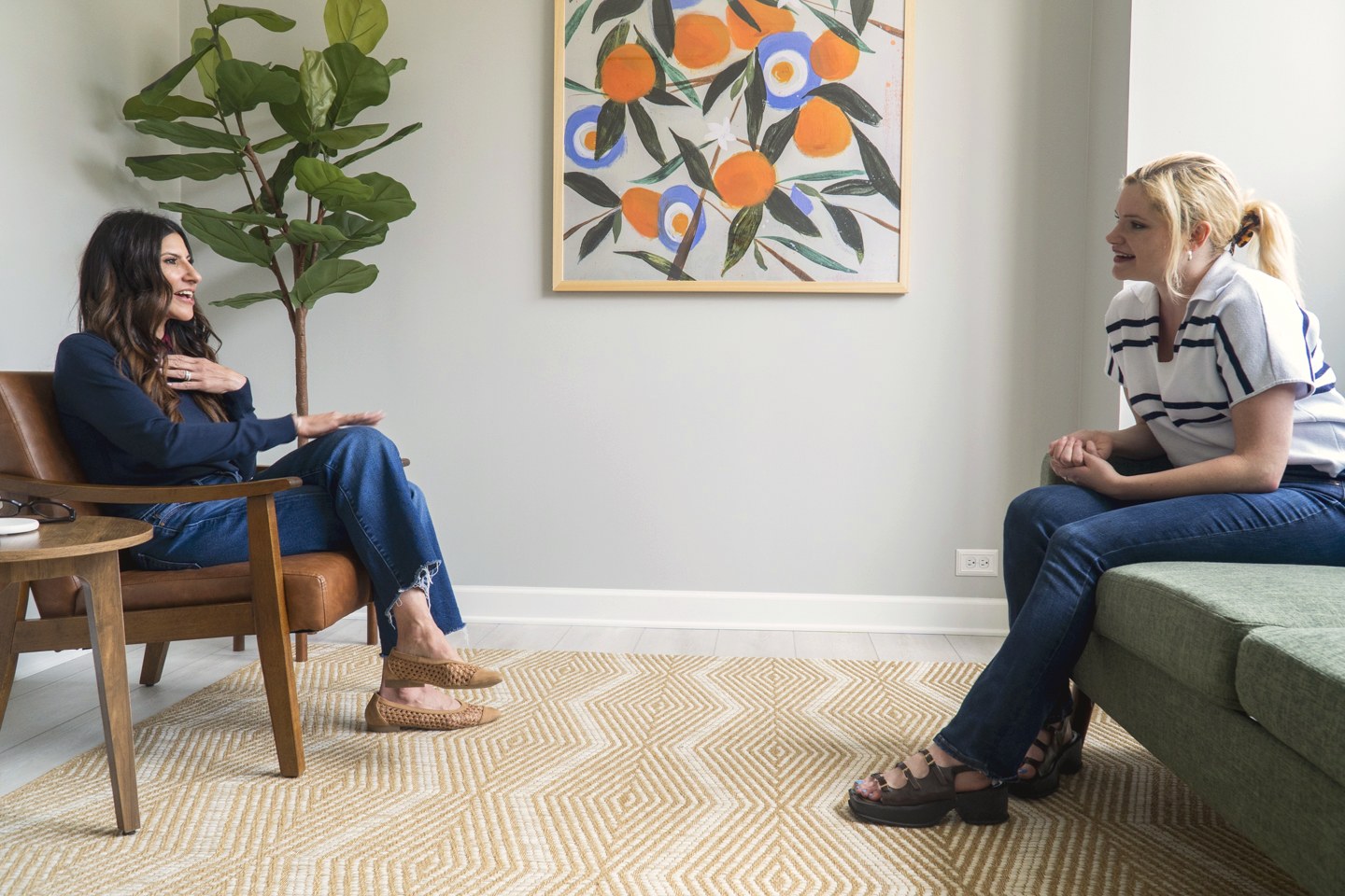 Two therapists in conversation, one in a leather armchair and one on a green sofa, beneath orange-fruit artwork in our therapy office