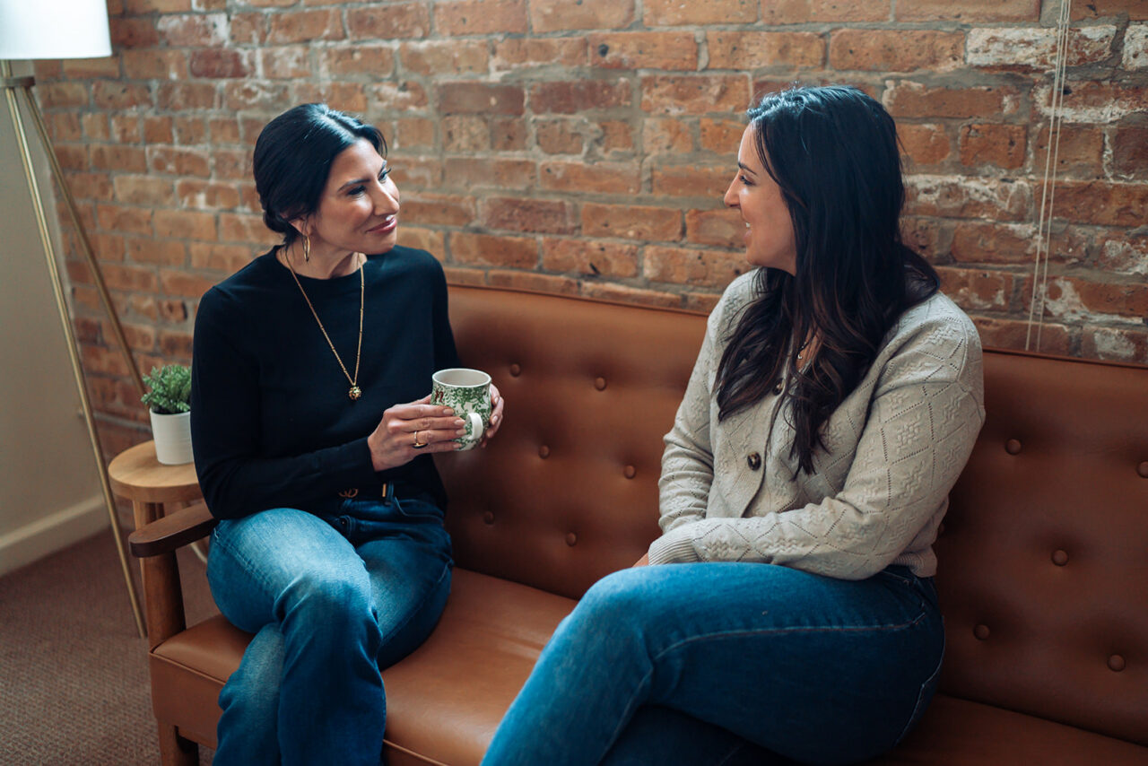 Two women talk and smile on a leather sofa in a warm, brick-walled therapy office