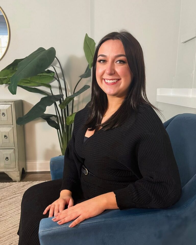 Kat Urrutia, AMFT, smiling in a blue armchair beside a large plant